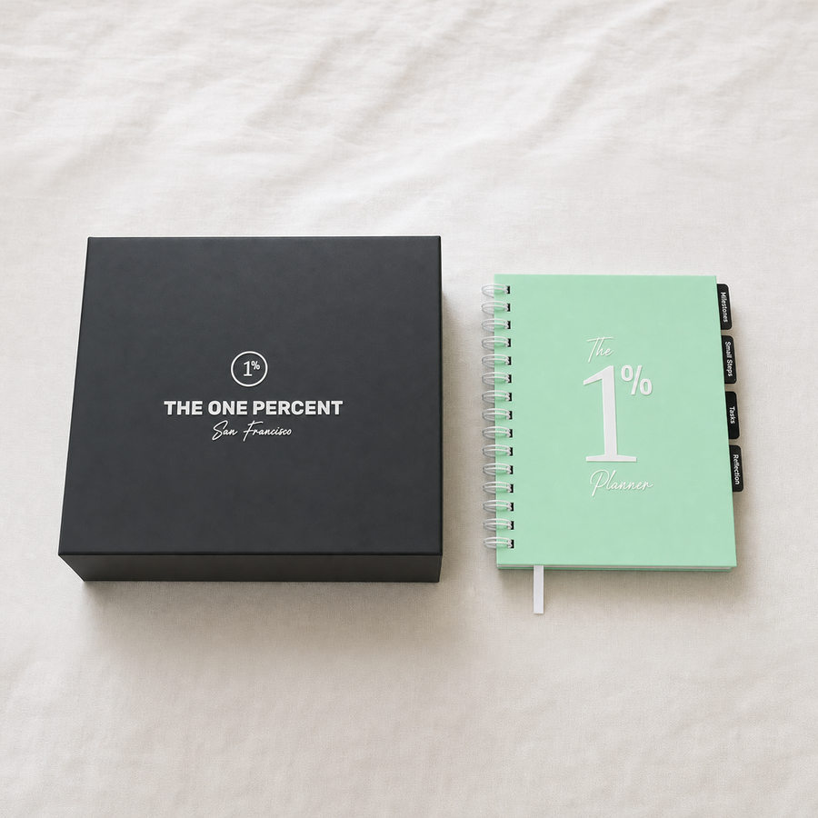 A mint green spiral-bound planner labeled 'The 1% Planner' sits next to a black presentation box printed with 'THE ONE PERCENT San Francisco' on a white fabric background.
